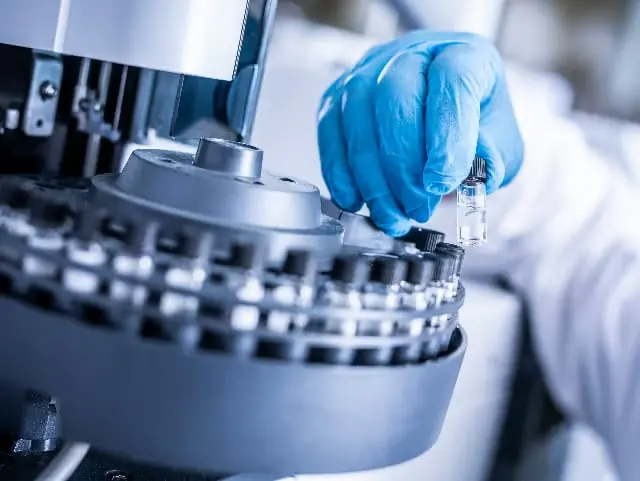 Gloved hand placing a sample vial into an automated laboratory analyzer